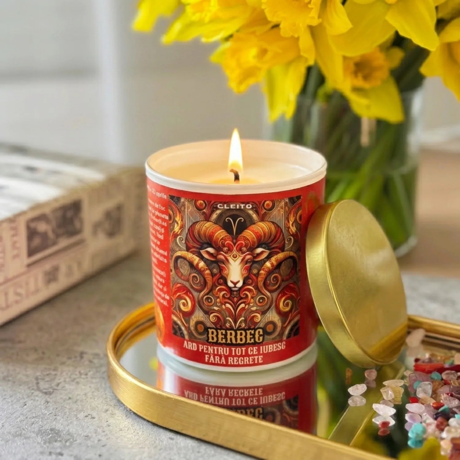 Lumânare parfumată Cleito Zodia Berbec aprinsă, cu flacără vie, plasată într-un decor cu flori, pe o tavita cu oglinda. Etichetă roșie detaliată cu ilustrație de berbec și textul „Ard pentru tot ce iubesc fără regrete”. Atmosferă placuta.